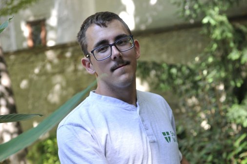 Ein junger Mann mit Brille, Schnauzer und weißem Stadt Wien-T-Shirt steht vor einer bewachsenen Mauer
